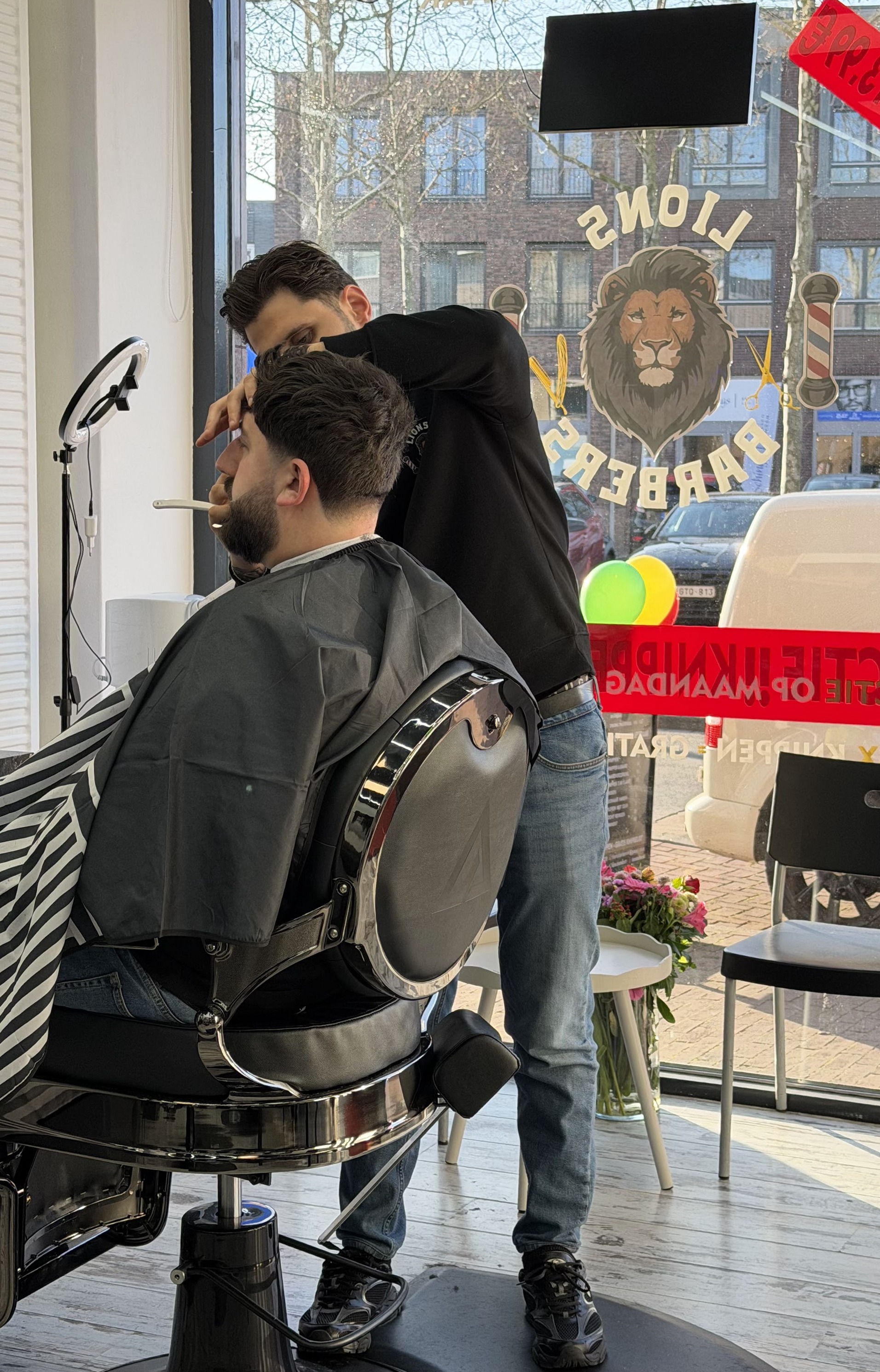 De kapper aan het werk bij het raam, onder de grote leeuw van Lions Barbers op het glas