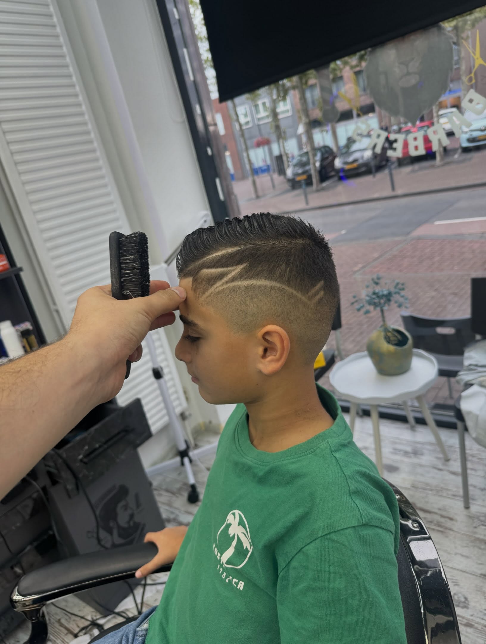 Jongen krijgt een zigzag hair design geschoren, met de straat achter het raam